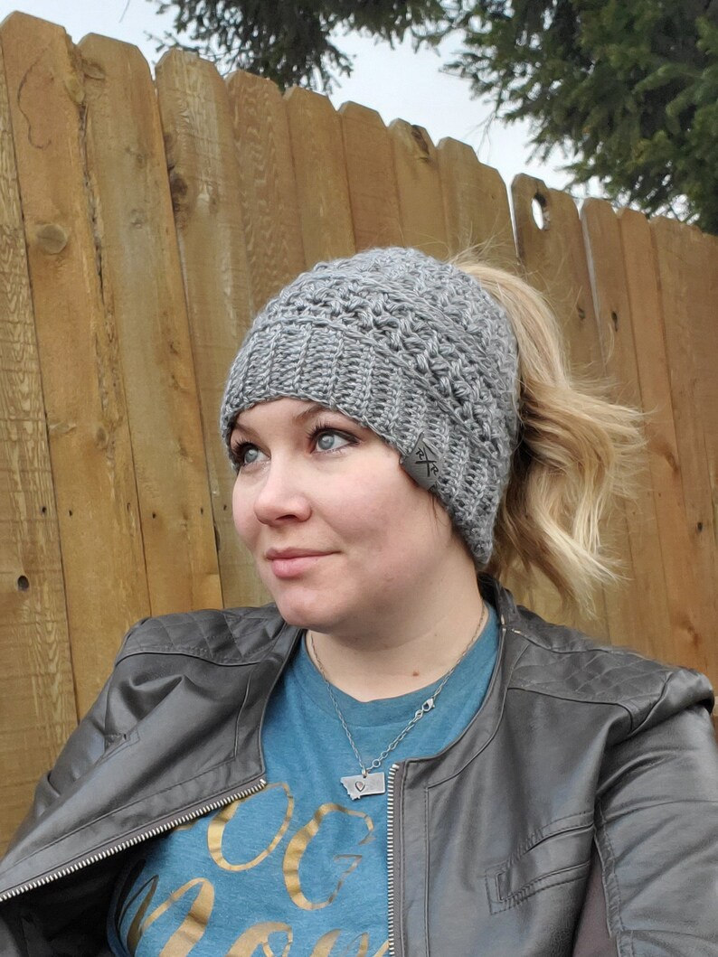 May include: A gray crocheted beanie with a ponytail opening. The hat has a textured pattern and a small tag on the side. The person is wearing a gray leather jacket and a blue shirt.