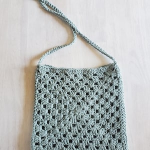 Puede incluir: Un bolso de ganchillo azul claro, de forma cuadrada, con una correa larga. El bolso presenta un detallado patrón calado, y la correa también está tejida a ganchillo. El bolso está diseñado para llevarse al hombro.