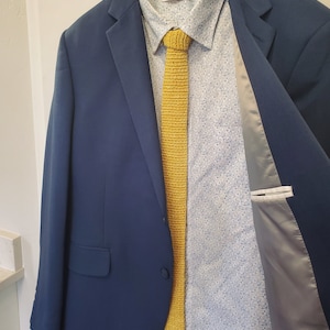 May include: A navy blue blazer with a yellow crocheted tie and a patterned button-down shirt. The blazer is buttoned and the tie is knotted. The shirt has a floral pattern.
