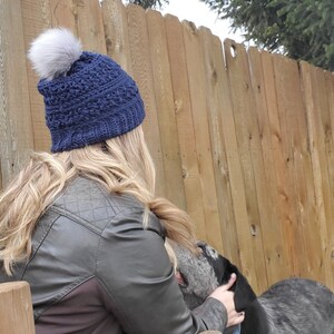 May include: A navy blue knit hat with a gray faux fur pom-pom. The hat has a textured pattern and is worn by a person with long blonde hair. The person is wearing a dark green quilted jacket. The background includes a wooden fence.
