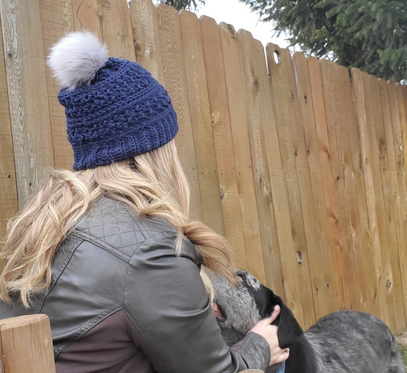 May include: A navy blue knit hat with a gray faux fur pom-pom. The hat has a textured pattern and is worn by a person with long blonde hair. The person is wearing a dark green quilted jacket. The background includes a wooden fence.