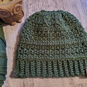 May include: A green crocheted beanie hat with a ribbed brim and a textured stitch pattern. A small brown leather tag with the initials "RR" is attached. The hat is shown next to a spool of green yarn and a decorative owl figurine.