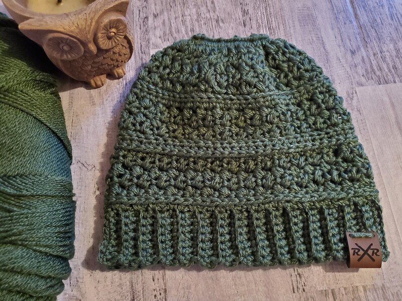 May include: A green crocheted beanie hat with a ribbed brim and a textured stitch pattern. A small brown leather tag with the initials "RR" is attached. The hat is shown next to a spool of green yarn and a decorative owl figurine.