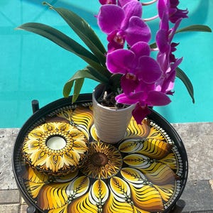 May include: A round black metal side table with a decorative sunflower design in yellow, brown, and white. A potted purple orchid and a candle holder with a sunflower design sit on the table.