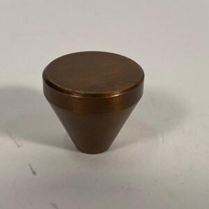 Könnte beinhalten: Ein konischer Schrankknopf in Bronzefarbe mit einer flachen, runden Oberseite.
