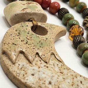 May include: A handcrafted necklace featuring a large, speckled ceramic pendant with a crescent moon shape and a series of colorful beads. The beads include red, green, and amber tones, with brass-colored accents.