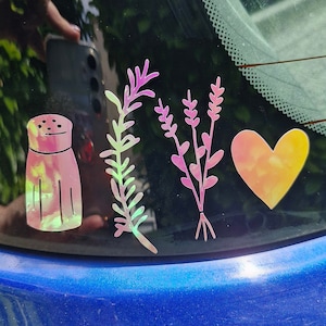 May include: A car window with four colourful decals: a salt shaker, a sprig of rosemary, a bunch of lavender, and a heart. The decals are iridescent and have a rainbow effect.