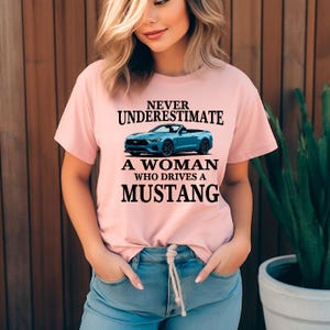 May include: A pale pink t-shirt with the slogan "Never Underestimate A Woman Who Drives a Mustang." The design includes a blue convertible Mustang. The model is wearing blue jeans.