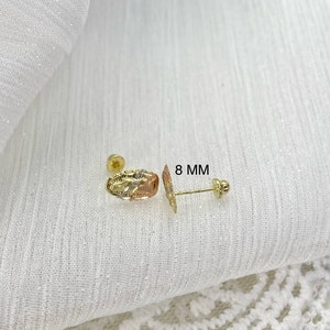 Op de afbeelding: Een paar gouden oorstekers. E&eacute;n oorbel heeft een ovale vorm met een textuurontwerp en driekleurig goud. De andere oorbel is een pin met een kleine ronde stud. De tekst "8 MM" is zichtbaar.