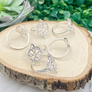 May include: Five silver rings with a Celtic knot design and clear rhinestones. The rings are arranged on a wooden surface.