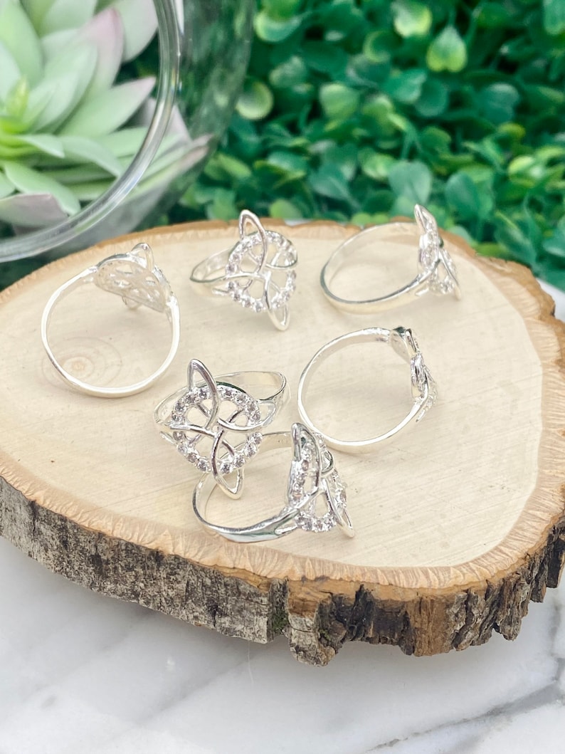 May include: Five silver rings with a Celtic knot design and clear rhinestones. The rings are arranged on a wooden surface.