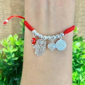 May include: Red braided bracelet with silver beads and charms. Charms include a Hamsa hand, an evil eye, a pearl, and a coin. The bracelet is worn on a wrist.