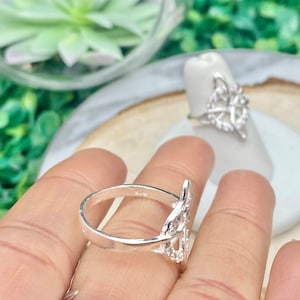 May include: A silver ring with a delicate, intricate design featuring a knot pattern. The ring is worn on a finger and is a statement piece of jewelry.