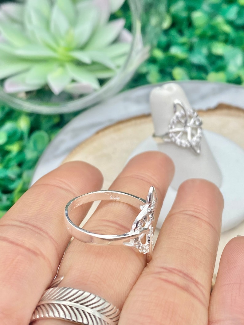 May include: A silver ring with a delicate, intricate design featuring a knot pattern. The ring is worn on a finger and is a statement piece of jewelry.