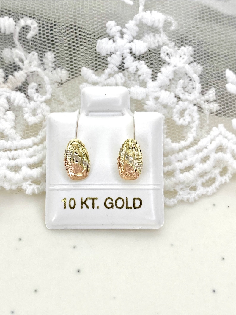 Op de afbeelding: Een paar goudkleurige oorstekers met een gedetailleerd ontwerp, gepresenteerd op een witte standaard. De oorbellen zijn gemarkeerd met "10 KT. GOLD". De achtergrond is een wit oppervlak met een kantpatroon.