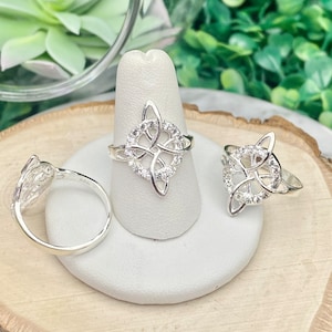 May include: Three silver rings with a Celtic knot design. The rings are on display on a white ring holder. The rings are set with small clear stones.