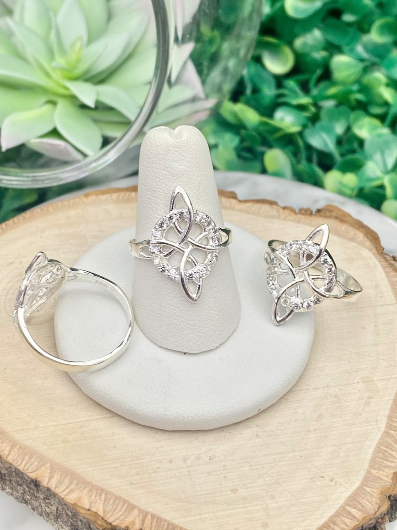May include: Three silver rings with a Celtic knot design. The rings are on display on a white ring holder. The rings are set with small clear stones.