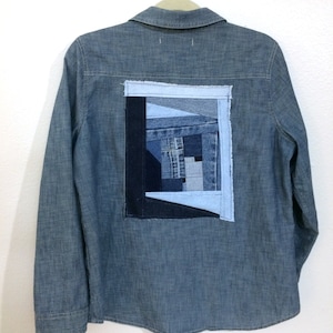 May include: A blue denim shirt with a patchwork design on the back. The patchwork is made of different shades of blue denim and is arranged in a geometric pattern.