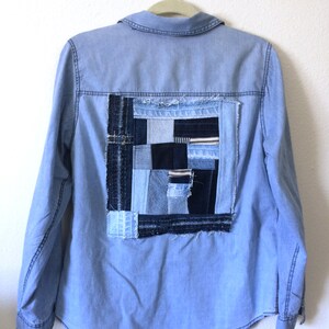 Könnte beinhalten: Ein hellblaues Jeanshemd mit einem großen Patchwork-Quadrat auf der Rückseite. Das Patchwork besteht aus verschiedenen Blautönen von Jeans und schwarzem Stoff.