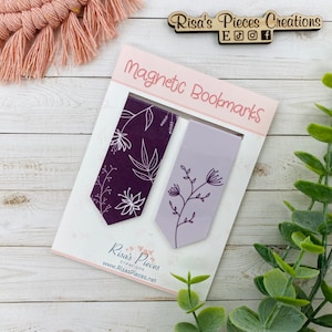 May include: Two magnetic bookmarks with floral designs. The bookmark on the left is purple with white floral designs. The bookmark on the right is light purple with dark purple floral designs. The bookmarks are displayed on a white wooden surface.