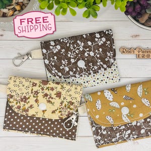 May include: Three fabric coin purses with snap closures and key rings. The purses feature floral and patterned designs in brown, yellow, and white. The purses are made of fabric and have a button closure.