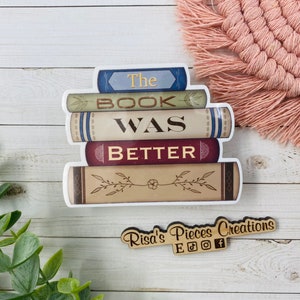 May include: A stack of books with the text "The Book Was Better" on a white background. The books are decorated with floral designs and have a vintage look. The image also includes a small wooden sign with the text "Risa's Pieces Creations" and social media icons.