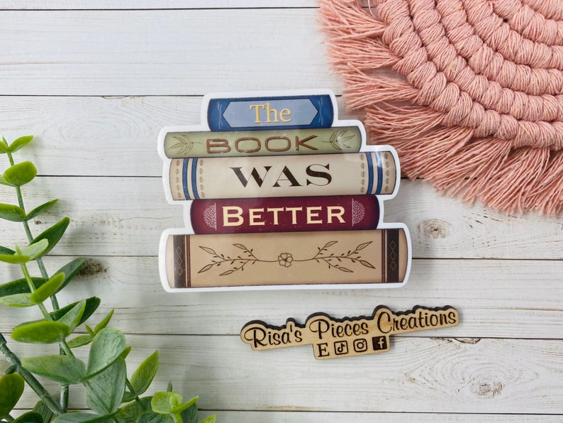 May include: A stack of books with the text "The Book Was Better" on a white background. The books are decorated with floral designs and have a vintage look. The image also includes a small wooden sign with the text "Risa's Pieces Creations" and social media icons.