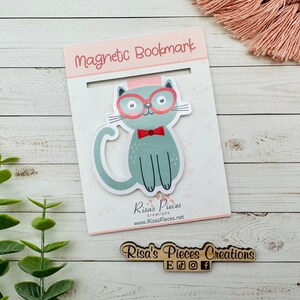 May include: A gray cat with red glasses and a red bow tie on a white magnetic bookmark. The bookmark says "Magnetic Bookmark" and "Risa's Pieces Creations" with a website address.