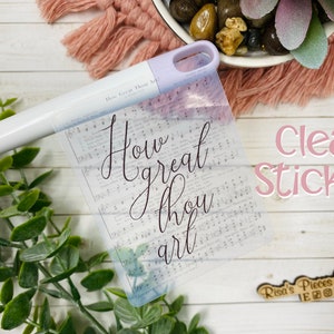 May include: Clear sticker with the text "How great thou art" printed on it. The sticker is on a white wooden surface with a green plant and a pink fuzzy surface in the background.