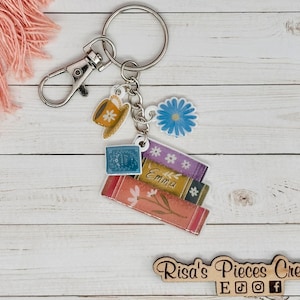 May include: A silver keychain with a stack of books, a teacup, a flower, and a charm. The books have a floral design and the top book says "Emma". The keychain is on a white wooden surface.