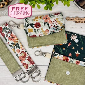 May include: A collection of floral print accessories, including a wallet, coin purse, and two keychains. The items feature a mix of red, white, and yellow flowers on a white background, with green accents. The keychains have silver hardware.