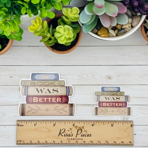 May include: Two stackable stickers with the text "The Book Was Better" on them. The stickers are designed to look like a stack of books with a vintage aesthetic. The stickers are on a white wooden surface.