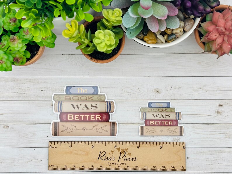 May include: Two stackable stickers with the text "The Book Was Better" on them. The stickers are designed to look like a stack of books with a vintage aesthetic. The stickers are on a white wooden surface.