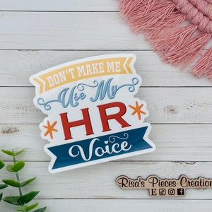 May include: A white sticker with the text "Don't Make Me Use My HR Voice" in colorful letters. The sticker has a blue, red, and yellow color scheme. The sticker is on a white wooden background.