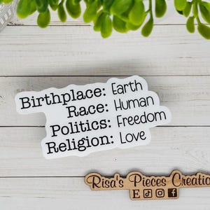May include: A white sticker with black text that reads: Birthplace: Earth, Race: Human, Politics: Freedom, Religion: Love. The sticker is on a white wooden surface, with a small wooden sign below it.