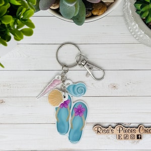 May include: A blue flip flop keychain with a seashell, a seashell, and a pink seashell charm. The keychain has a silver key ring and a silver lobster clasp. The keychain is on a white wooden surface. Risa's Pieces Creations Etsy, Facebook, Instagram, and Twitter social media icons are on a wooden sign.
