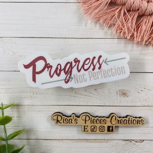 May include: White sticker with the text "Progress Not Perfection" in maroon lettering with a grey arrow pointing to the right. The sticker is on a white wooden surface. Below the sticker is a small wooden sign with the text "Risa's Pieces Creations" in black lettering and social media icons.