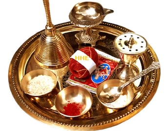May include: A brass thali set featuring a handbell, diya, incense stick holder, spoon, and bowls. The set includes roli and mauli. The thali is decorated with a beaded border. HHE is written on a red packet.