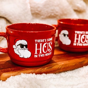 May include: Two red ceramic mugs with white text and a cartoon Santa Claus design. The text on the mugs reads "There's some HOS in this house".