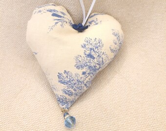 Op de afbeelding: Een wit hartvormig kussen met een blauw bloemenpatroon en een blauwe kraal die eronder hangt. Het kussen heeft een witte lintlus om op te hangen.