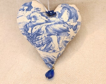 Op de afbeelding: Een blauwe en witte hartvormige stoffen ornament met een blauwe glazen kraal die aan de onderkant hangt. De ornament heeft een witte lus om op te hangen.