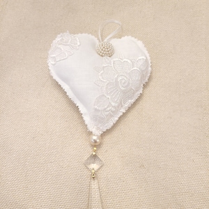 May include: White heart-shaped cushion with lace trim and a pearl button. A crystal teardrop hangs from the bottom.