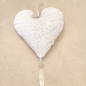 Könnte beinhalten: Weißes herzförmiges Ornament mit Spitze und gepunktetem Stoff. Es hat eine Perlenverzierung und eine Schleife. Ein klarer Kristalltropfen hängt von unten. Geeignet für Hochzeiten oder besondere Anlässe.
