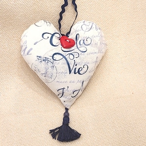 May include: A white heart-shaped cushion with a black tassel and a red button. The cushion has a French phrase printed on it that says "C'est la Vie".