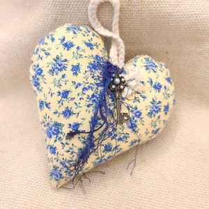 Könnte beinhalten: Ein gelbes und blaues, herzförmiges Kissen mit Blumenmuster und einem Schlüsselanhänger. Das Kissen hat eine weiße Schlaufe aus Seil zum Aufhängen.
