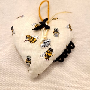 Puede incluir: Un adorno de tela en forma de corazón con un patrón de abejas y un pequeño amuleto de abeja. El adorno está hecho de tela beige con un borde negro y una cinta negra.