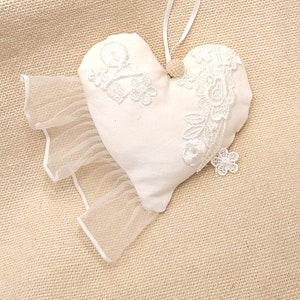 May include: A white heart-shaped pillow with lace trim and a white ribbon loop for hanging. The pillow is on a beige burlap background.