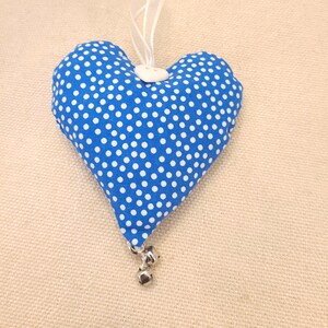 May include: A blue heart-shaped cushion with white polka dots and a white button. The cushion has a white ribbon loop and silver bells attached.