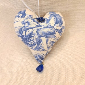 May include: A blue and white heart-shaped fabric ornament with a blue glass bead hanging from the bottom. The ornament has a white loop for hanging.