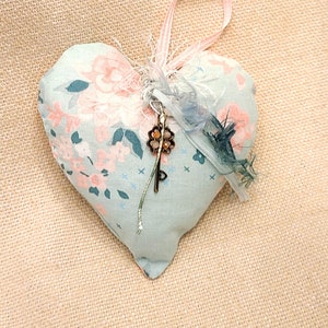 Könnte beinhalten: Ein hellblaues herzförmiges Kissen mit einem Blumenmuster und einem Schlüsselanhänger. Das Kissen hat ein rosafarbenes Band und ist mit Federn verziert.